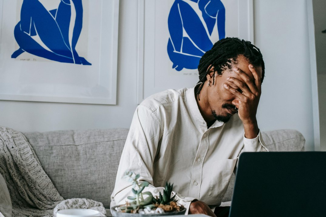 A man on a couch holds his hand over his face in front of a laptop