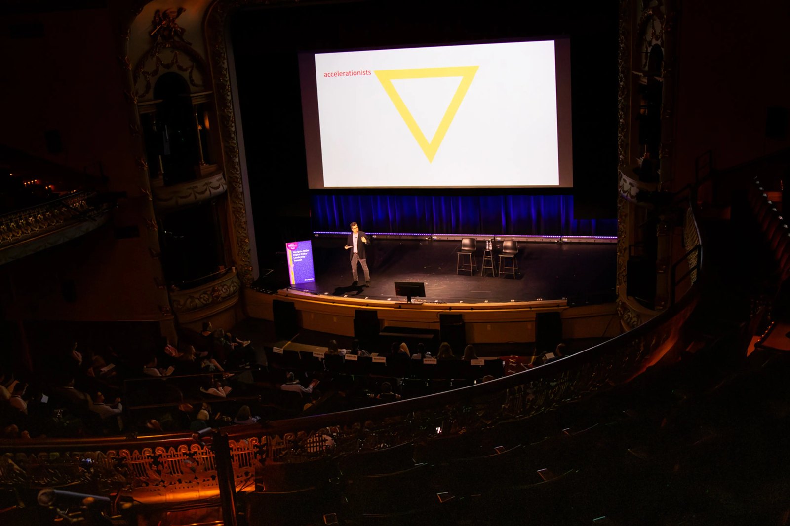 The Music Hall in Portsmouth seen from the balcony: a speaker on a dark stage beneath a large screen showing a yellow triangle labeled accelerationists, with the audience below