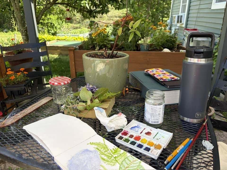 An outdoor table set for painting: an open watercolor sketchbook with a hydrangea and fern, a paint box, brushes, and potted flowers behind